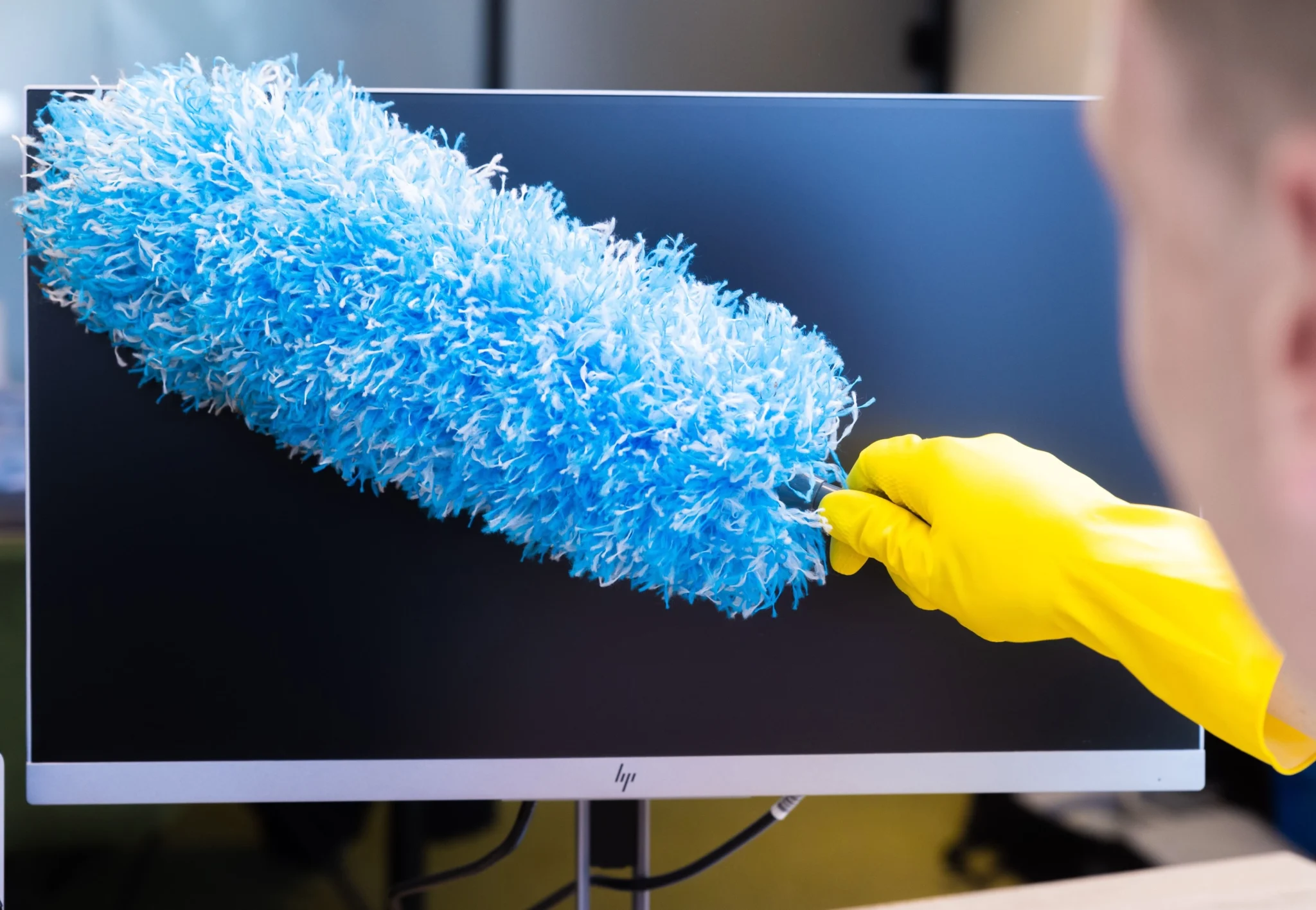 een hand met een gele handschoen en een blauwe stoffer in zijn hand die een beeldscherm schoonmaakt